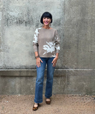 Woman wearing a beige sweater with white floral patterns and blue jeans standing against a gray wall.