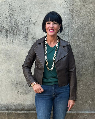 Woman wearing a brown leather jacket, green top, and blue jeans standing against a concrete wall.