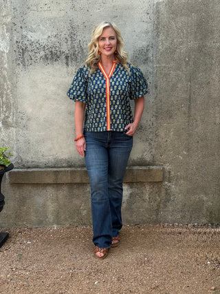 Woman wearing a patterned blouse and jeans standing against a textured wall.