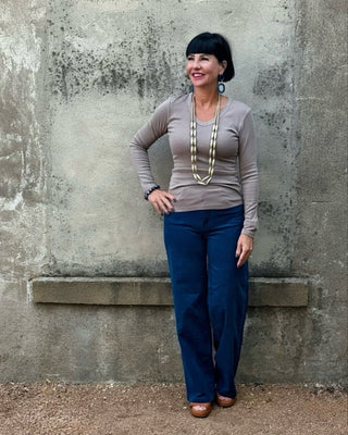 Woman standing against a textured wall wearing a gray long-sleeve top and blue pants.
