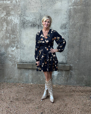 Woman in a floral dress and silver boots standing against a concrete wall.