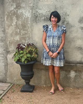 Woman in a blue patterned dress standing next to a stone wall and planter with greenery.