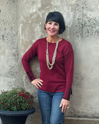 Woman wearing a burgundy top and blue jeans standing against a textured wall with a potted plant.