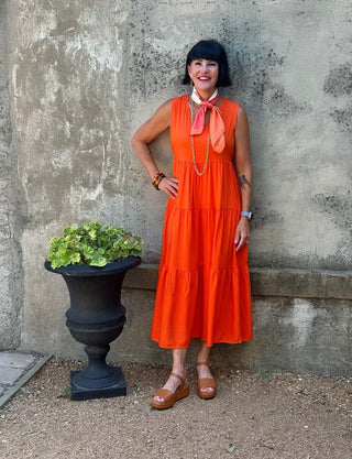 A woman with a black bob haircut stands against a textured concrete wall wearing a sleeveless tiered orange midi dress with a coral neck scarf. She is wearing brown flat sandals and standing next to a black planter with green leaves.
