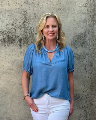 Woman wearing a blue blouse and white pants standing against a textured wall.