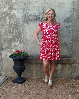 Woman in a red and white floral dress standing against a textured wall with a planter of flowers.