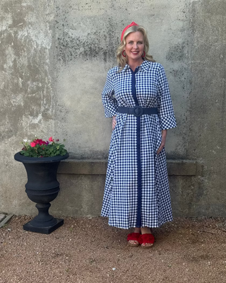 Woman in a blue checkered dress standing against a textured wall with a pot of flowers.