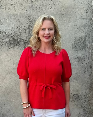 Woman in a red top and white pants standing next to a planter with flowers against a textured wall.