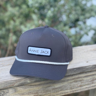 Gray cap with 'Annie Jack' logo on a wooden surface with blurred greenery in the background