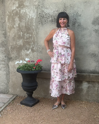 Woman in a floral dress standing next to a stone wall with a planter of flowers.