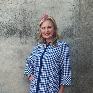 Woman in a blue checkered dress standing against a textured wall with a flower pot nearby.