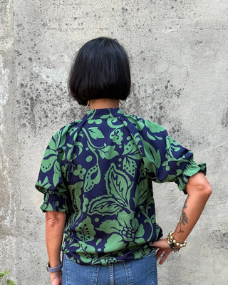 Person wearing a green and blue patterned shirt and blue jeans standing against a textured wall.