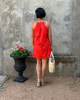 Woman in a red dress standing against a textured wall with a plant and bag nearby.