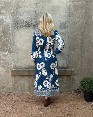 Person wearing a blue floral dress standing on a concrete surface with sand and plants in the background.