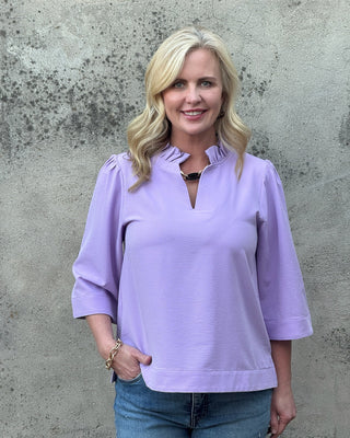 Woman wearing a light purple blouse and blue jeans against a textured gray wall.