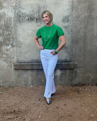 Woman in a green shirt and white pants standing against a textured wall.