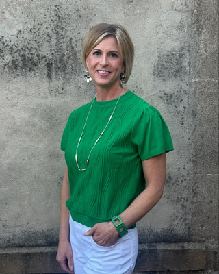 Woman wearing a green blouse and white pants standing against a textured wall.