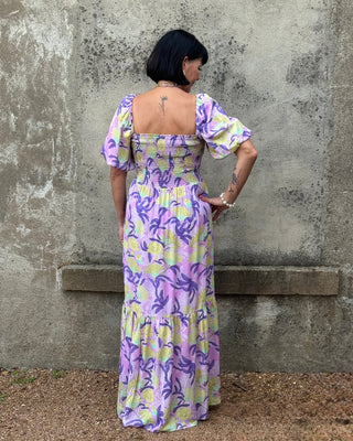 Woman wearing a floral dress with a pattern of purple and yellow standing against a textured wall.