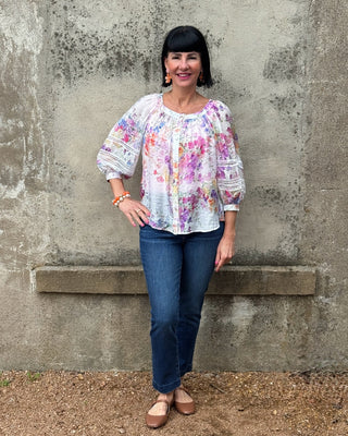 Woman wearing a colorful blouse and blue jeans standing against a textured wall.