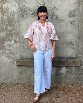 Woman wearing a floral blouse and white pants standing against a textured wall.
