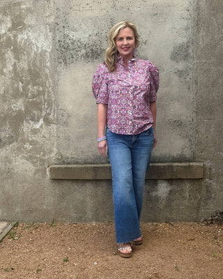 Woman in a patterned shirt and blue jeans standing against a textured wall.