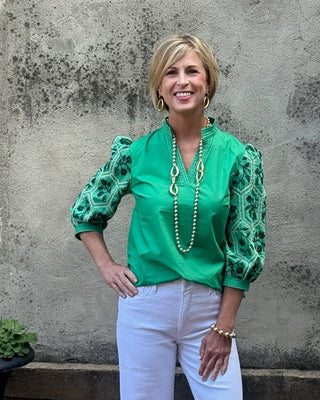 Woman in a green blouse and white pants standing against a textured wall with a pot of plants.