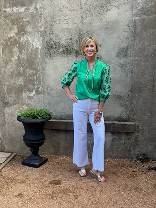 Woman in a green blouse and white pants standing against a textured wall.