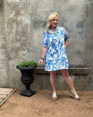 Woman in a blue floral dress standing next to a stone wall and black urn with greenery.