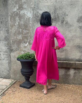 Person wearing a bright pink dress standing on a concrete surface with a plant nearby.