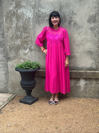 Woman in a bright pink dress standing against a textured wall with a potted plant nearby.