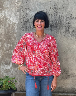 Woman in a red patterned blouse and blue jeans standing next to a potted plant against a textured wall.
