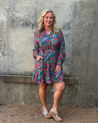 Woman wearing a colorful floral dress standing against a textured wall.