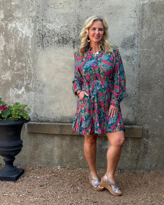 Woman in a colorful dress standing against a textured wall with a potted plant.