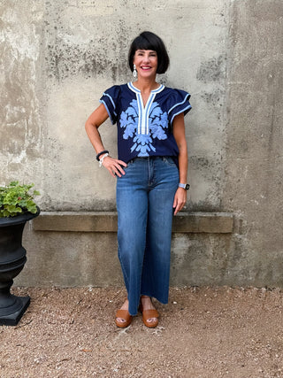 Woman wearing a blue floral top and jeans standing against a stone wall.