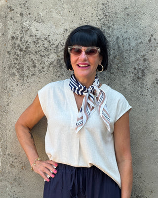 Woman wearing a white blouse and dark pants with a scarf, standing against a textured wall with flowers.