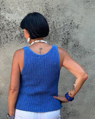 Woman in a blue sleeveless top and white pants standing next to a stone wall with a planter of flowers.
