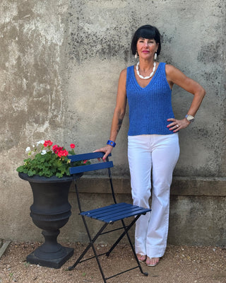 Woman in a blue sleeveless top and white pants standing next to a small table with flowers against a textured wall.
