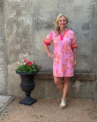 Woman in a pink floral dress standing on a concrete surface with a potted plant nearby.