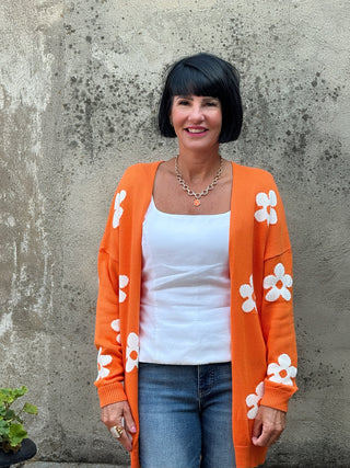 Woman wearing an orange cardigan with white floral patterns against a textured wall.