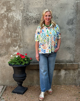 Woman in a floral shirt and jeans standing next to a potted plant against a textured wall.