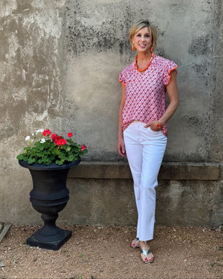 Woman in a pink top and white pants standing next to a potted plant against a textured wall.