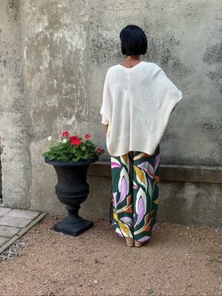 Person wearing a white top and colorful pants standing next to a potted plant against a textured wall.