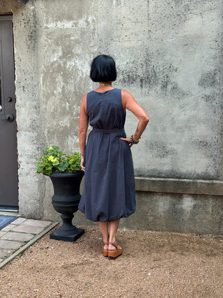 Woman in a blue dress standing in front of a textured wall with a planter nearby.