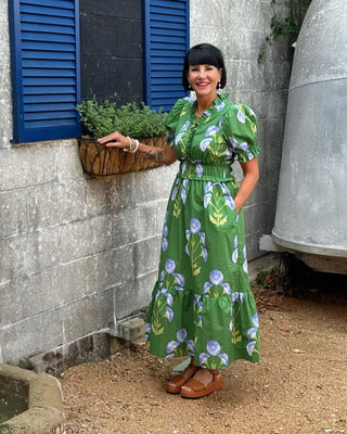 Woman in a green floral dress standing in front of a stone wall with blue shutters.