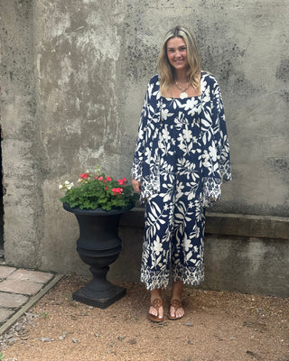 Woman in a blue floral dress standing next to a potted plant on a concrete surface.