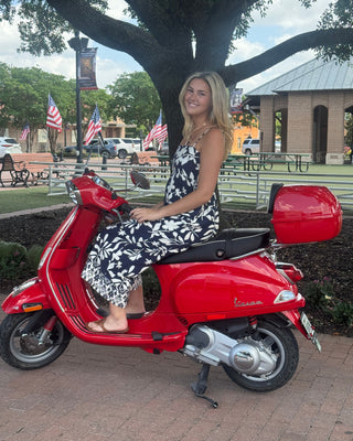 Woman sitting on a red scooter in an outdoor setting with flags and trees in the background