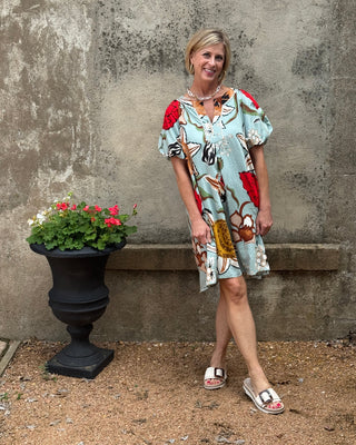 Woman in a floral dress standing next to a potted plant against a textured wall.