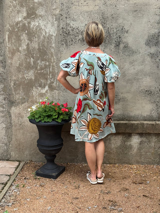 Child wearing a floral dress standing next to a potted plant against a textured wall.