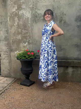 Woman in a blue and white dress standing next to a potted plant against a textured wall.