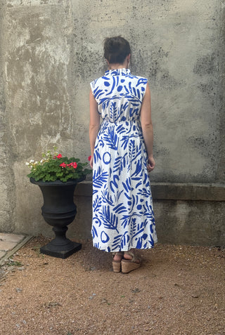 Person wearing a blue and white patterned dress standing in front of a textured wall with a planter of flowers.